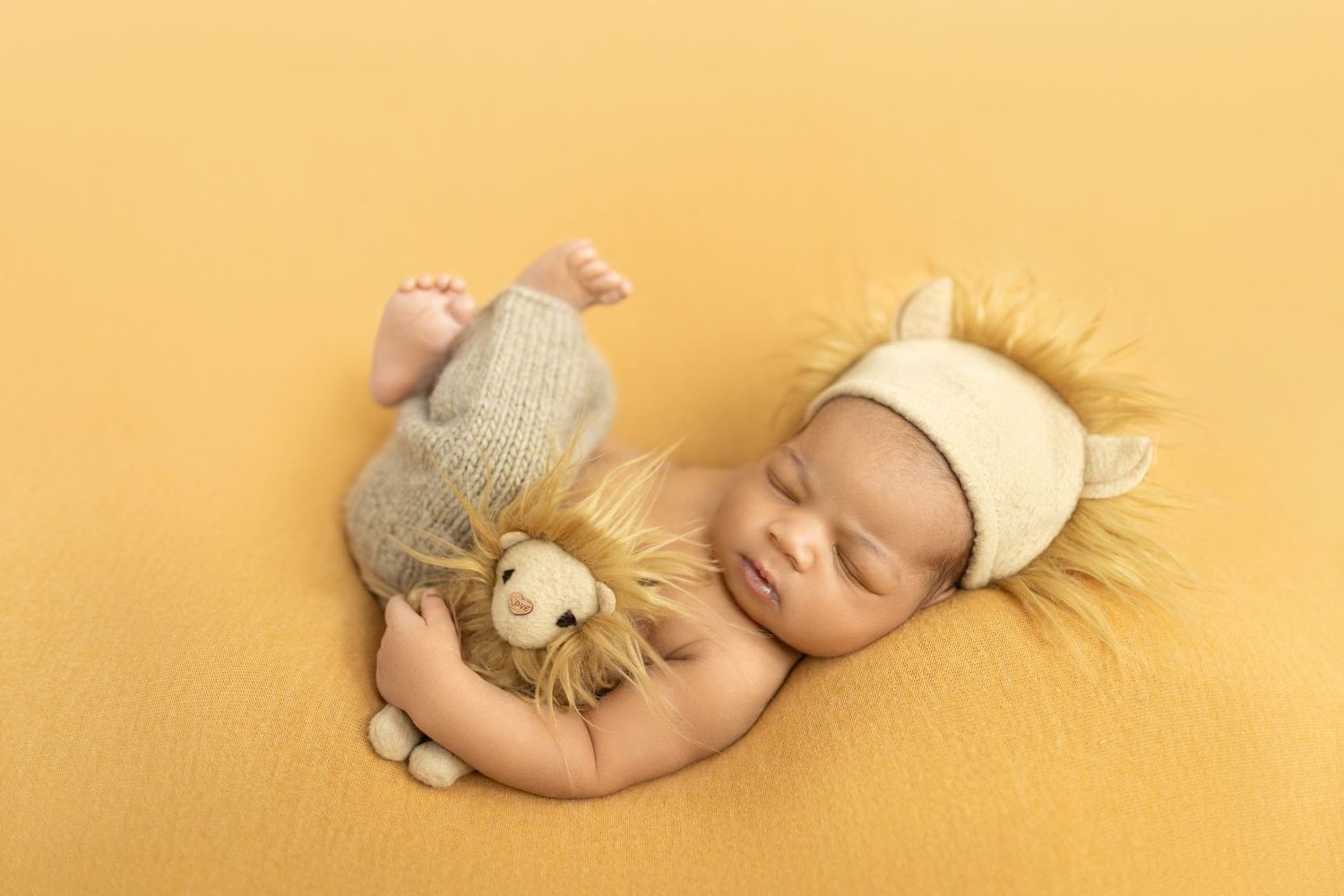 Newborn baby sleeping on yellow blanket wearing lion hat, cuddling a knitted lion stuffed animal toy.