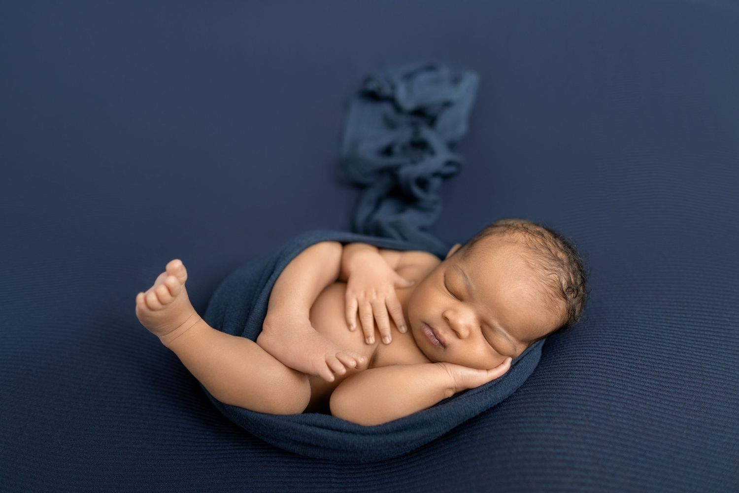 Newborn baby sleeping peacefully on a navy blue backdrop, wrapped in a soft blue cloth in a cozy posed position.