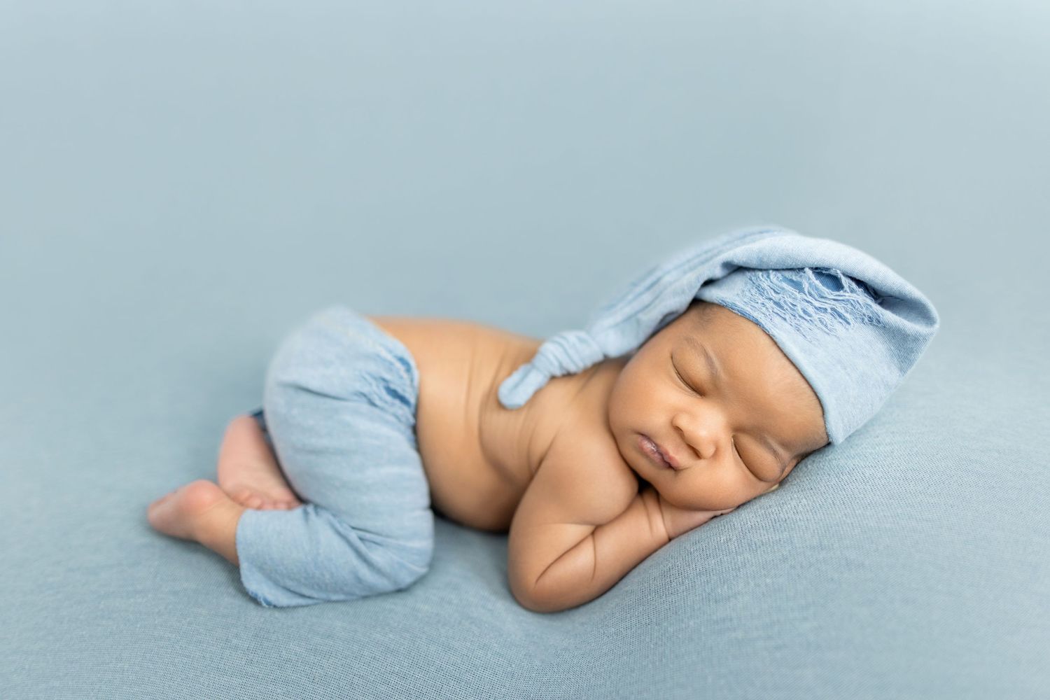 Sleeping newborn baby wearing a blue knit hat and pants, curled up peacefully on a soft blue blanket.