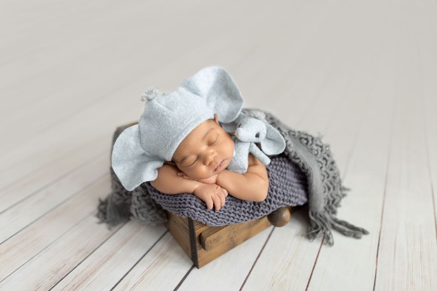 Sleeping newborn baby in gray knit hat with stuffed elephant toy in rustic wooden box with soft blanket.