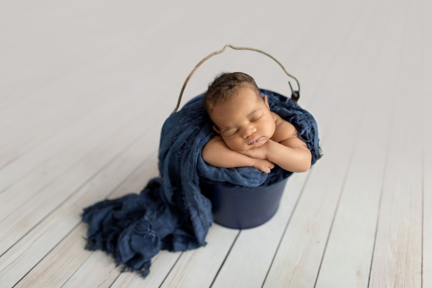 Sleeping newborn baby wrapped in navy blue cloth inside a dark blue bucket on white wood floor.