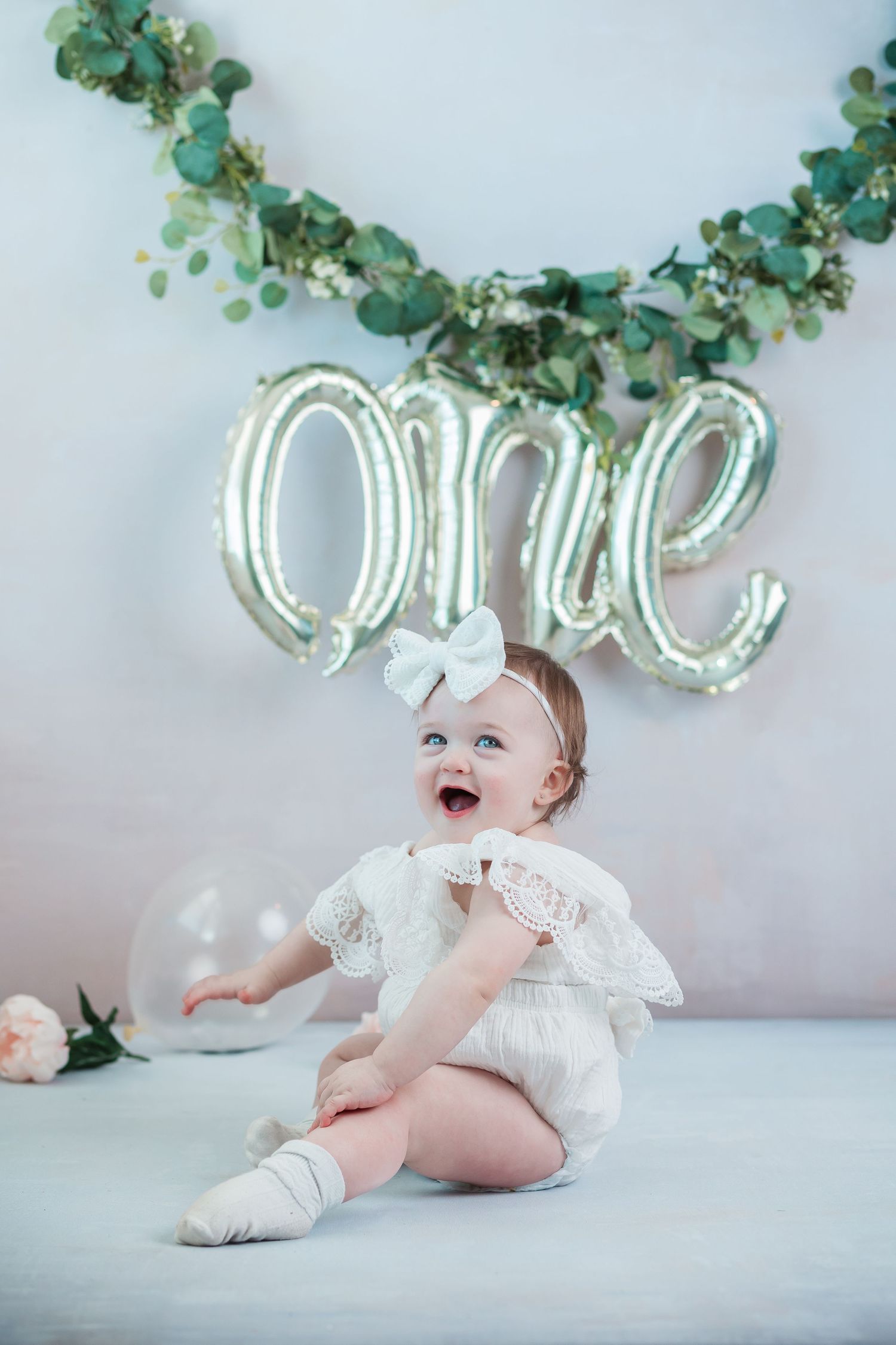 Smiling baby girl in white lace outfit celebrates first birthday with silver balloon and eucalyptus garland backdrop.