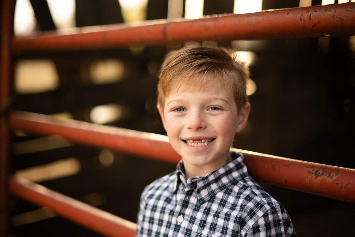 Smiling young boy with short hair wearing a plaid shirt, posing outdoors near red metal railings.