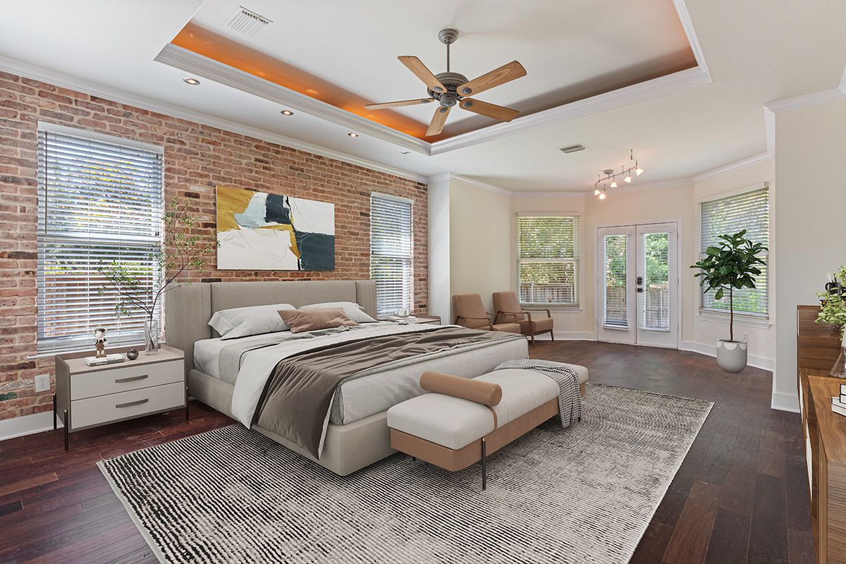 Spacious master bedroom with a tray ceiling, ceiling fan, exposed brick accent wall, king bed, bench, area rug and dark hardwood floors.