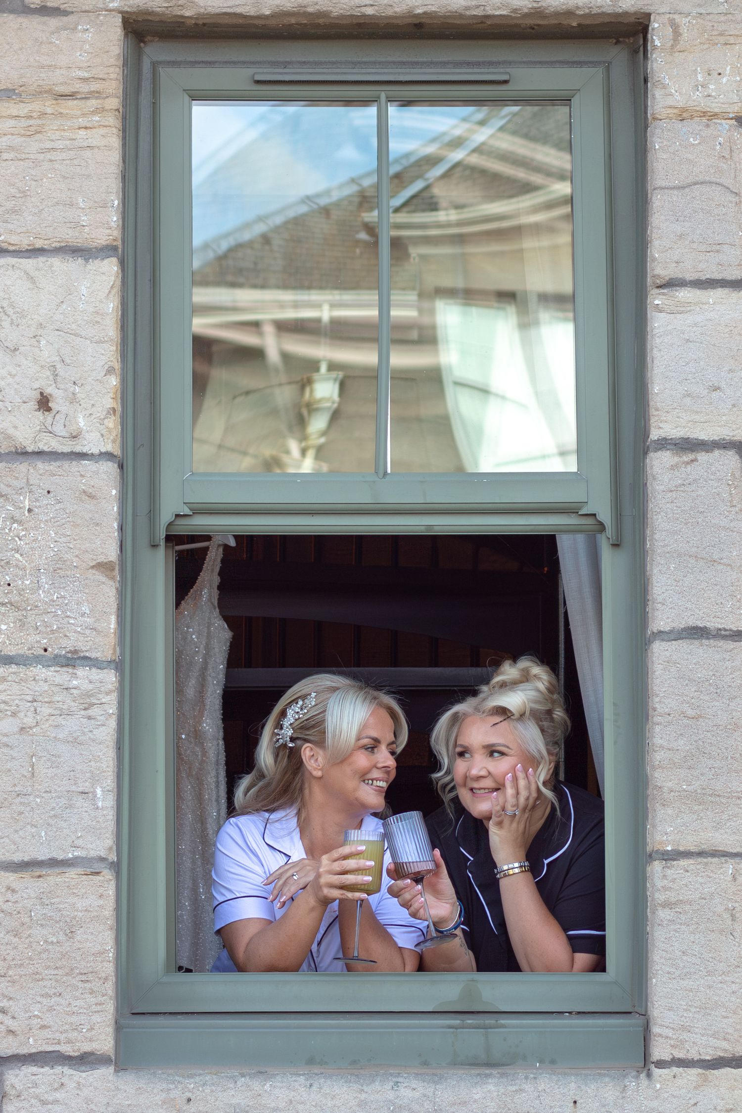 Two smiling women in a green-framed stone window hold drinks and laugh together, one with a floral hair accessory and one with an updo.
