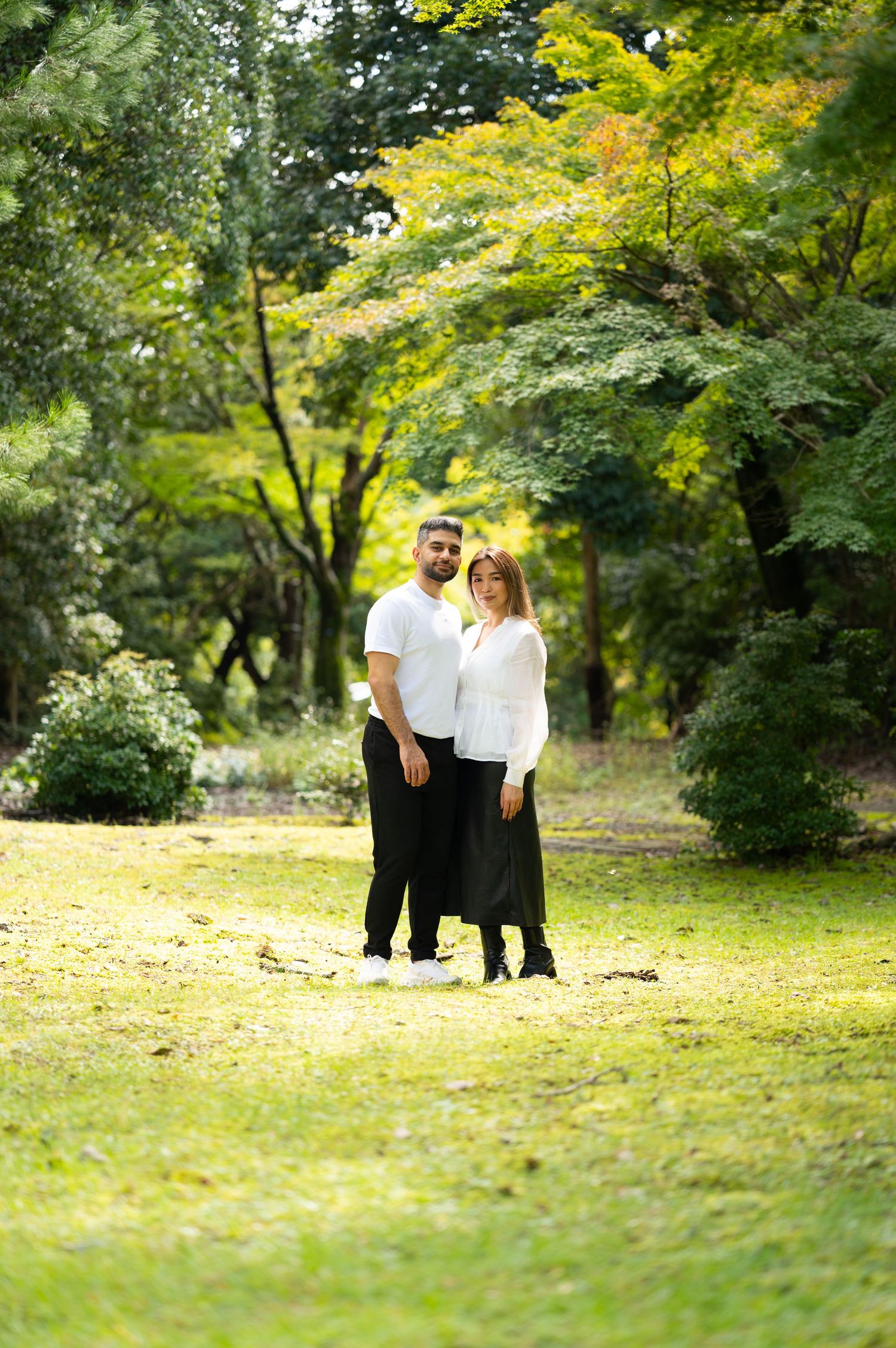 Couple in traditional Japanese kimono walking hand in hand along a sunlit garden path beneath lush green maple trees.