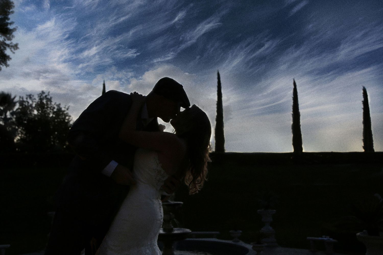 Silhouette of a bride and groom sharing a kiss against a dramatic blue evening sky with wispy clouds and tall cypress trees.