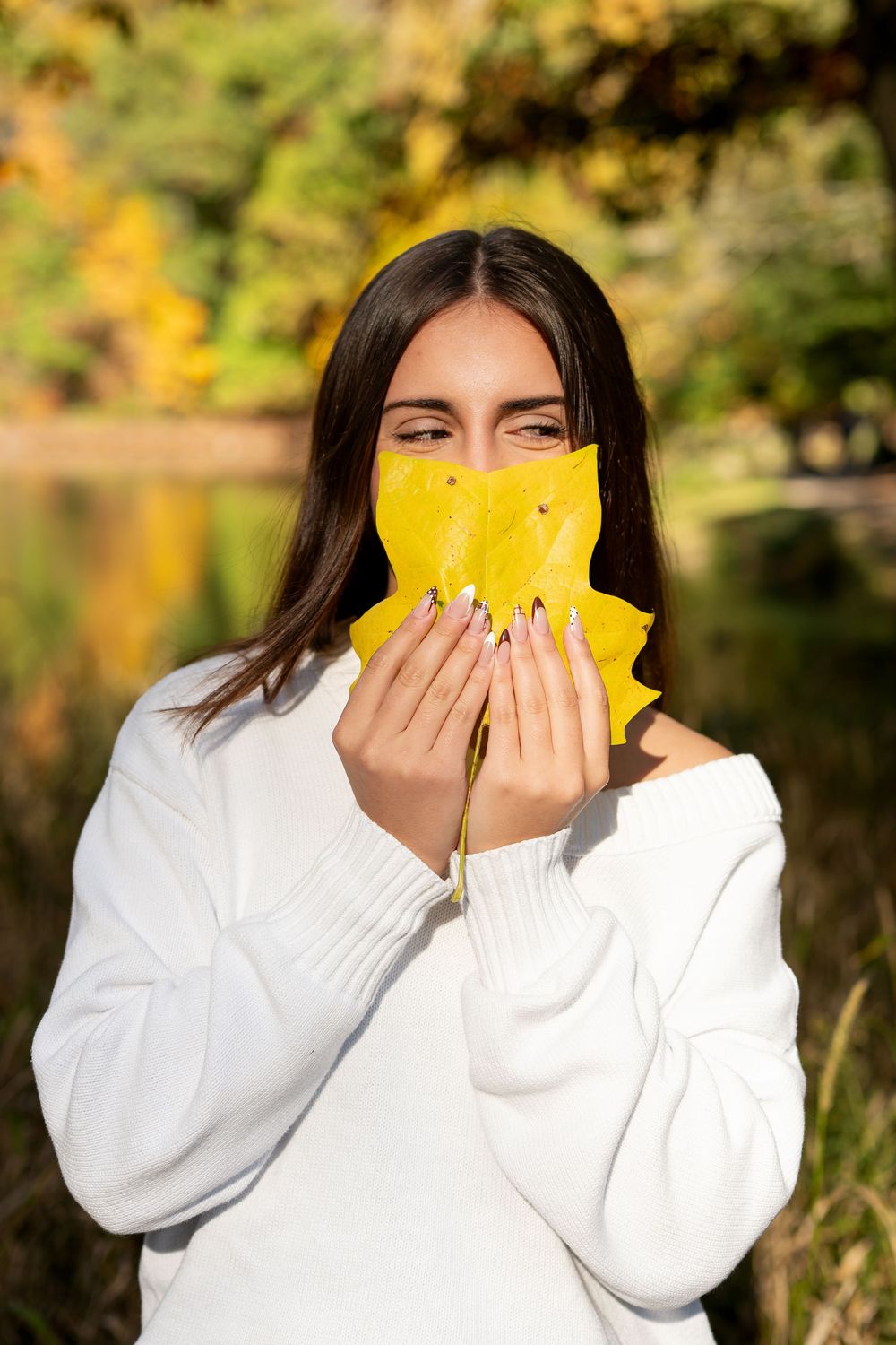 Young woman in a white knit sweater holds a yellow autumn leaf over her face by a lake with colorful fall foliage.