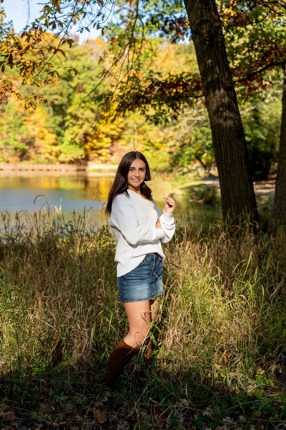 Young woman in a white sweater, denim shorts and brown boots poses in tall grass beside a lake with colorful autumn trees.