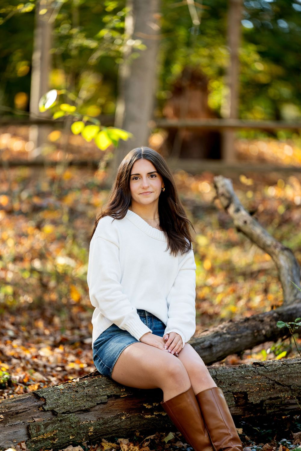 Smiling young woman in a cream sweater and denim shorts sits on a mossy log surrounded by fall foliage in the woods.