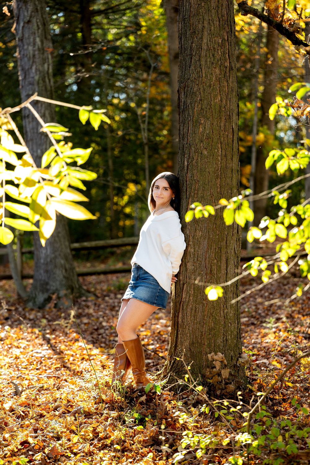 Young woman in a white sweater, denim skirt and brown boots leans against a tree in a sunlit autumn forest.