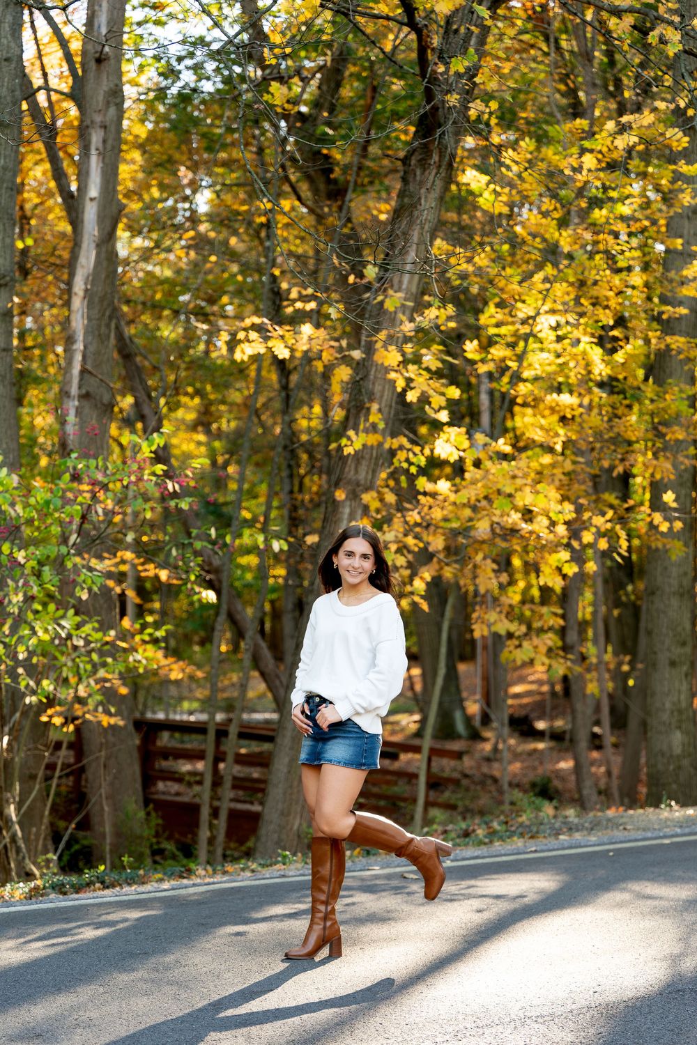 Young woman in a white sweater, denim shorts and tall brown boots poses on a tree-lined road surrounded by golden autumn foliage.