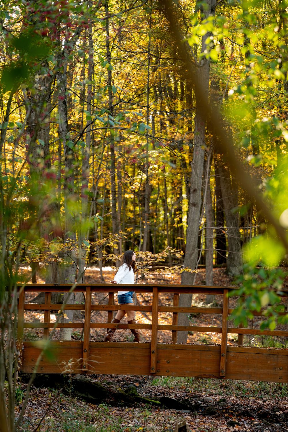 Girl in a white top and denim shorts walks across a wooden footbridge surrounded by golden fall foliage.