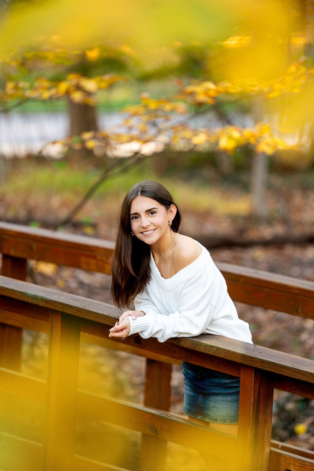 Smiling young woman with long dark hair in a white sweater leans on a wooden bridge railing surrounded by golden autumn leaves.