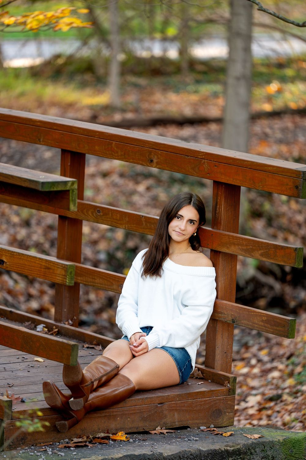 Young woman in a white off-shoulder sweater, denim shorts and brown boots sits on a wooden bridge surrounded by autumn leaves.