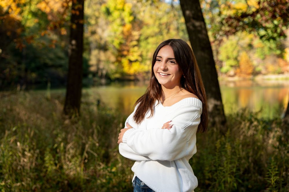 Smiling young woman with long dark hair and crossed arms in a white sweater stands in a grassy field near a lake with colorful autumn trees.