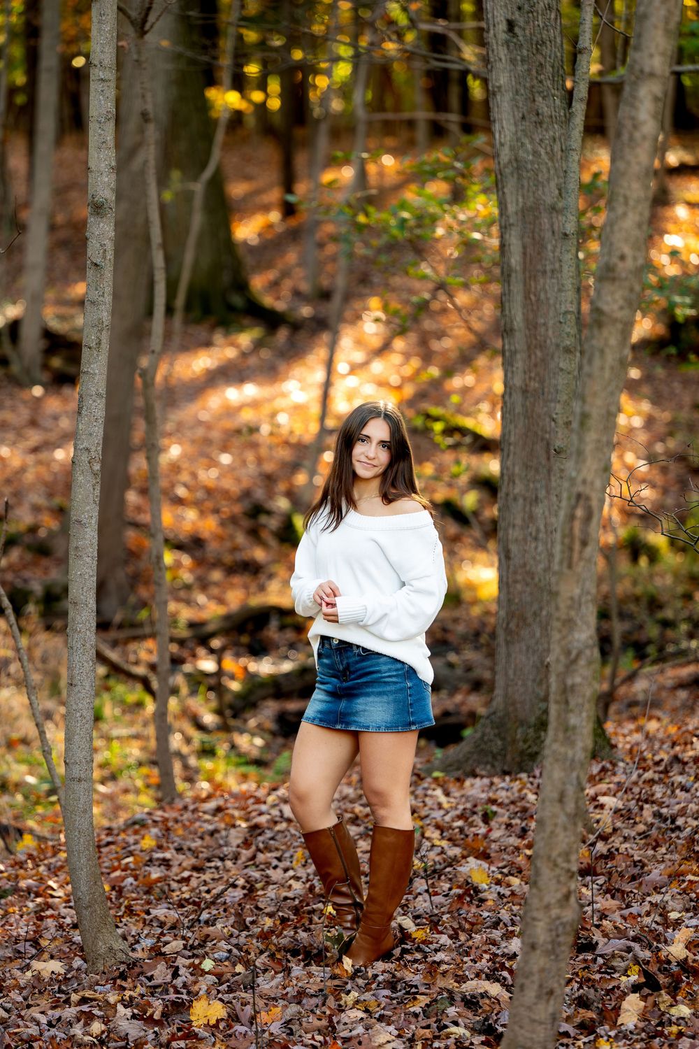 Young woman in a white off-shoulder sweater, denim skirt and brown boots poses among trees on a leaf-covered forest floor in autumn.