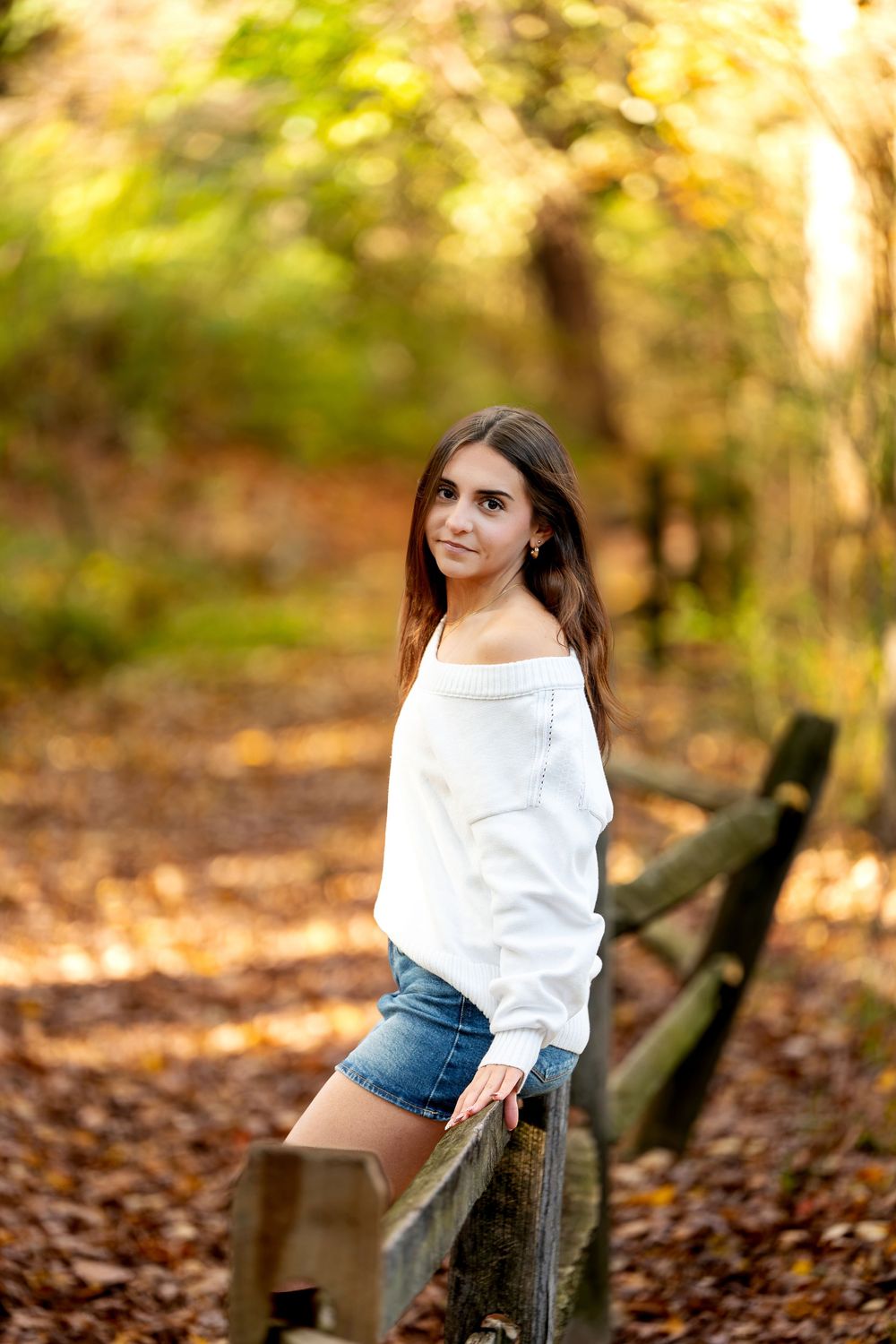 Young woman in a white off-shoulder sweater rests against a wooden fence beside a woodland path covered in autumn leaves.
