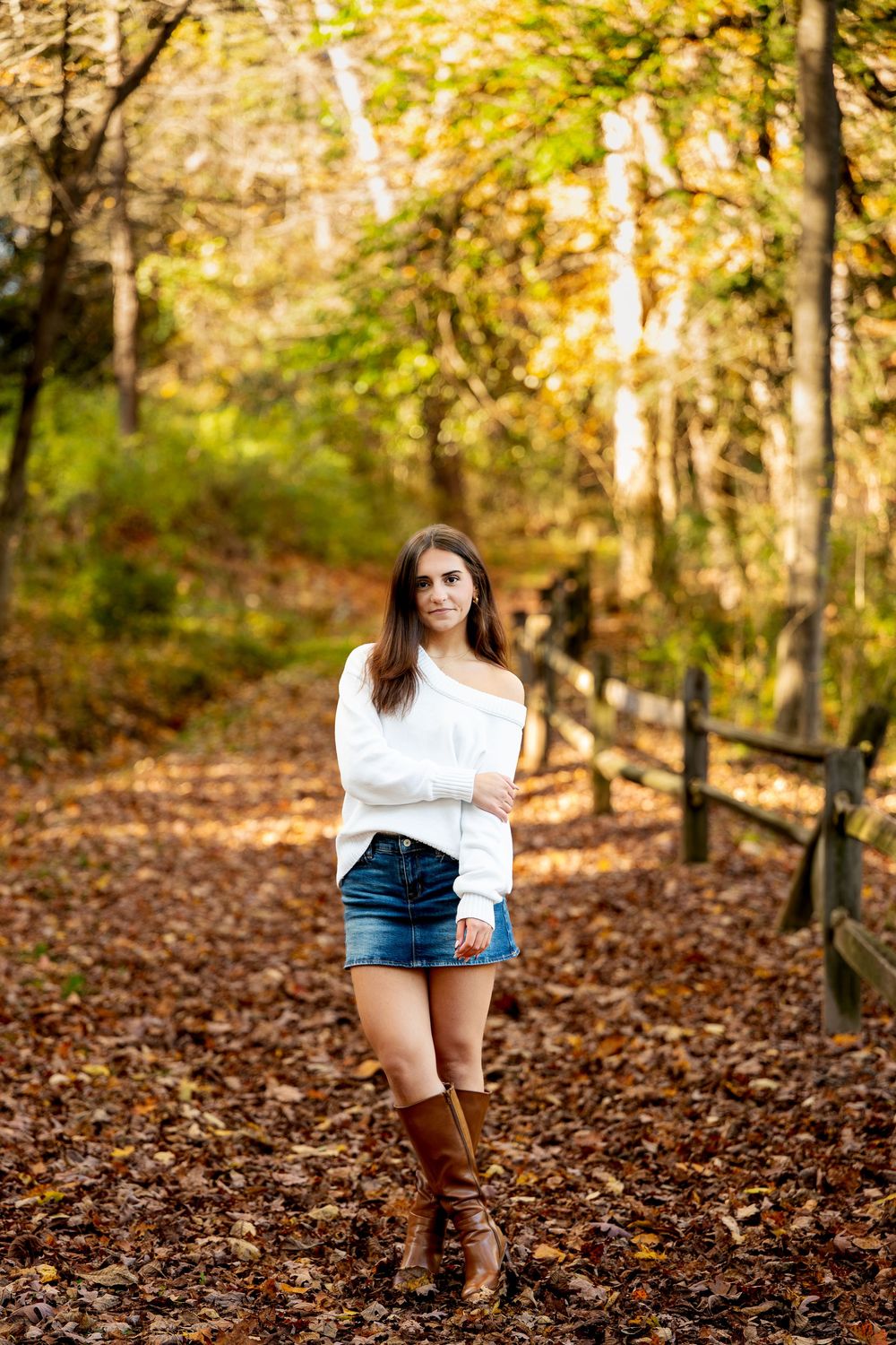 Young woman in an off-shoulder white sweater, denim skirt and brown boots stands on a leaf-covered trail in the fall.