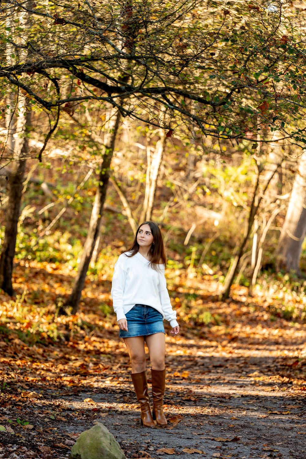 Young woman in a white sweater, denim skirt and tall brown boots walks along a leaf-covered forest path in warm autumn light.