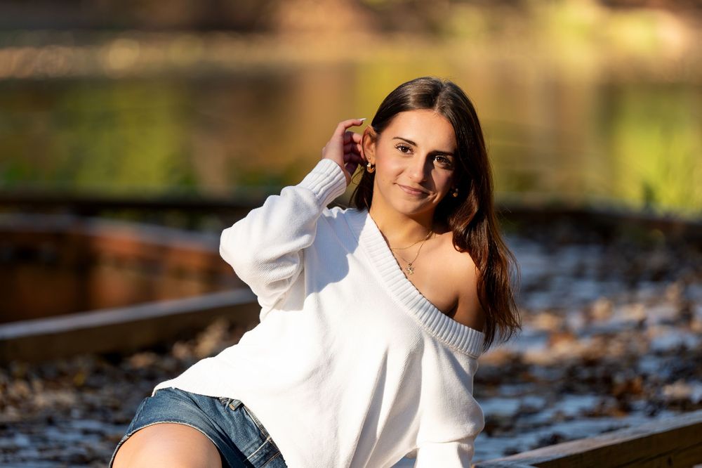 Young woman with long brown hair in an off-shoulder white sweater and denim shorts sits on railroad tracks, smiling in warm golden light.