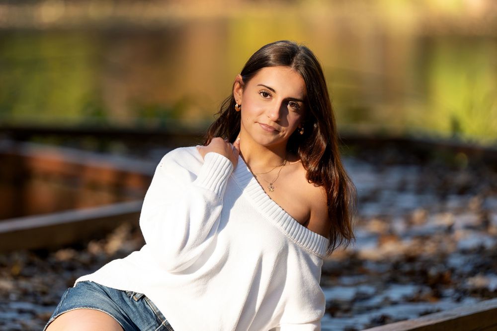 Young woman with long dark hair in an off-the-shoulder white sweater and denim shorts poses by railroad tracks in warm autumn light.