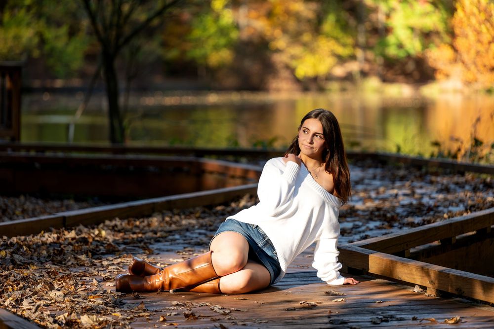 Young woman in a white sweater and boots sits on a wooden dock covered in fallen leaves beside a calm autumn lake.