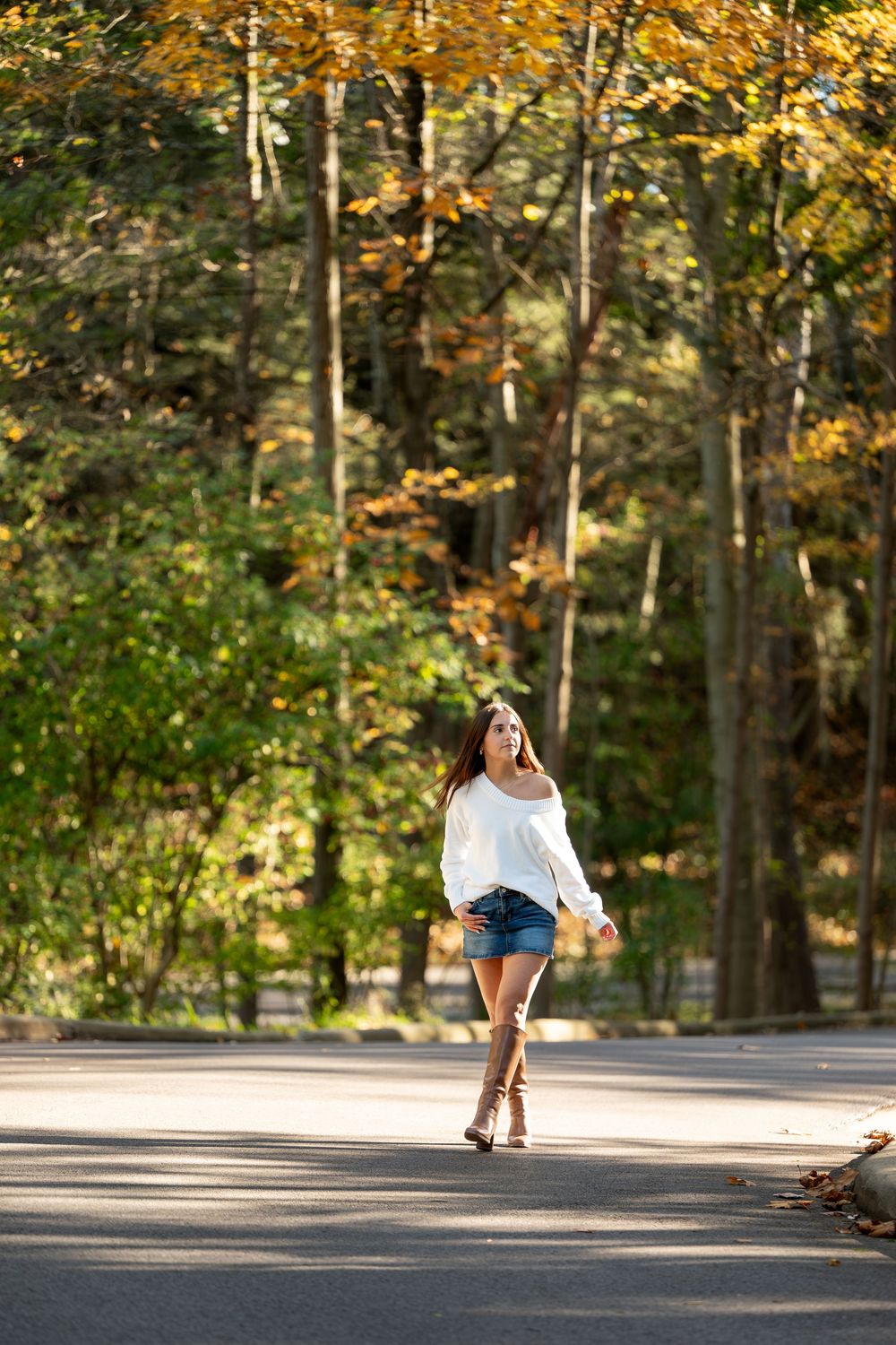 Young woman in an off-shoulder white top, denim shorts and tall boots walks along a sunlit road lined with autumn trees.