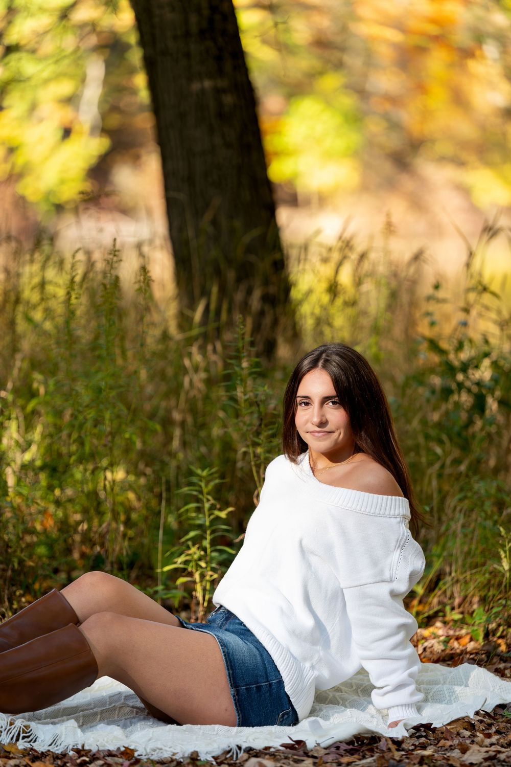 Young woman in a white off-shoulder sweater, denim shorts and brown boots sits on a blanket in an autumn forest with golden foliage.