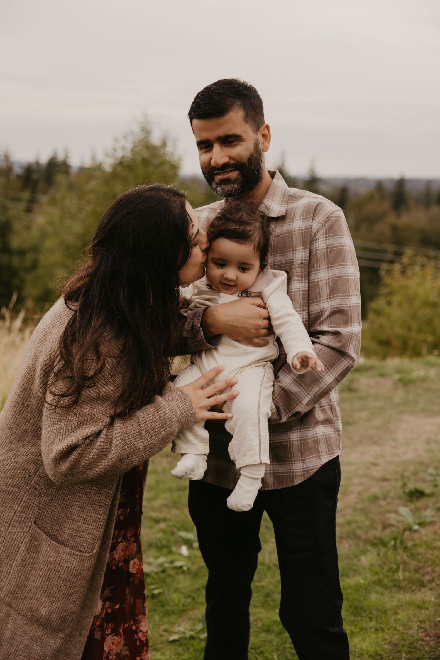 A woman in a knit cardigan kisses a smiling baby held by a bearded man in a plaid shirt, outdoors on a grassy hill with trees behind Monroe