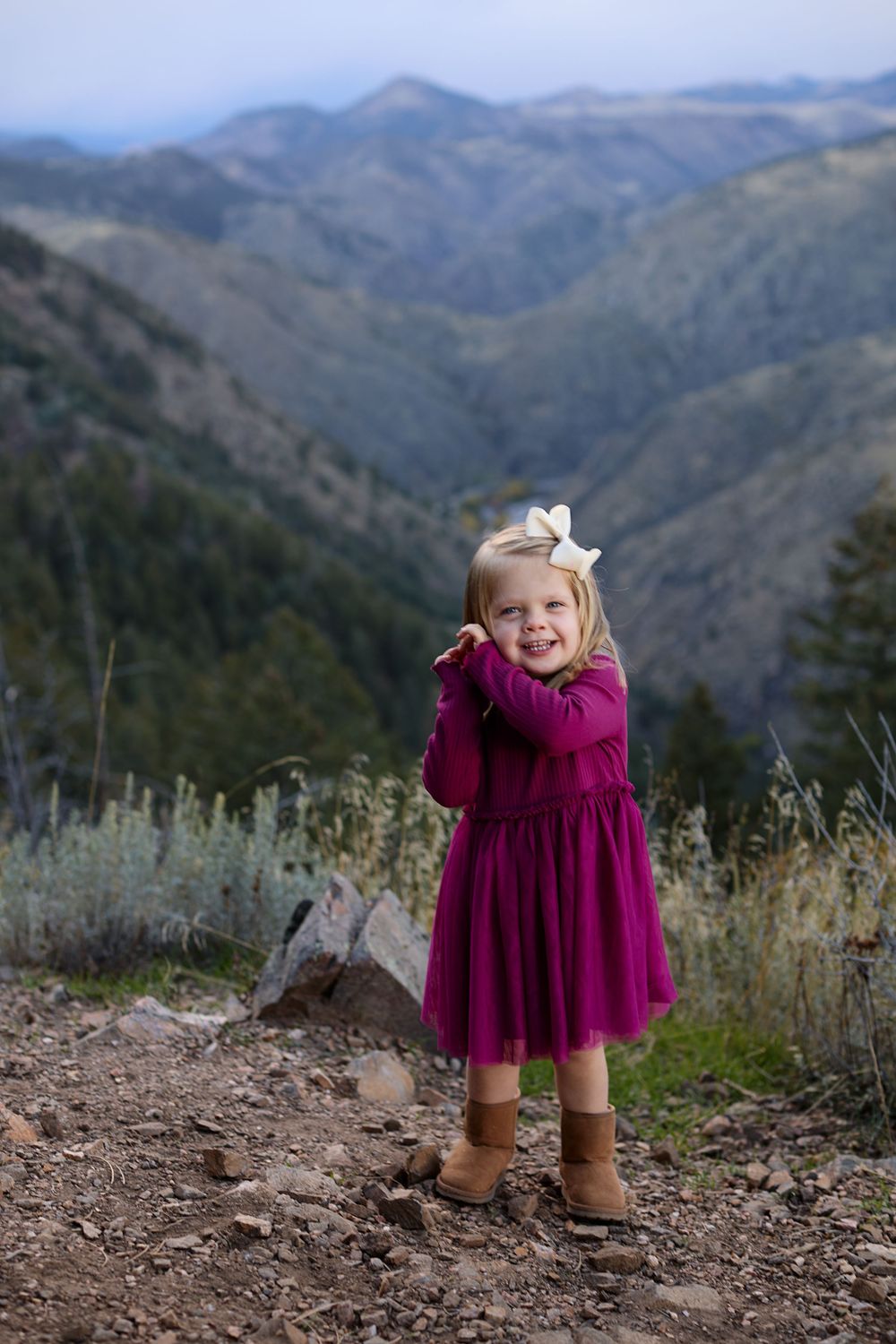 Smiling toddler girl in a magenta tulle dress, white hair bow and tan boots standing on a rocky mountain overlook with layered hills behind.