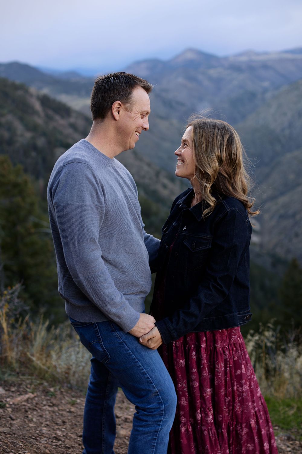 Smiling couple holding hands and facing each other on a mountain overlook with rolling hills in the background at dusk.