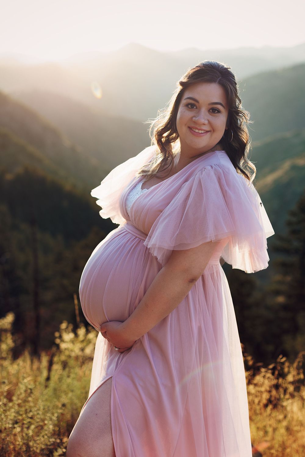 Smiling pregnant woman in a flowing pink tulle maternity gown cradling her belly on a mountain overlook at golden hour sunset.