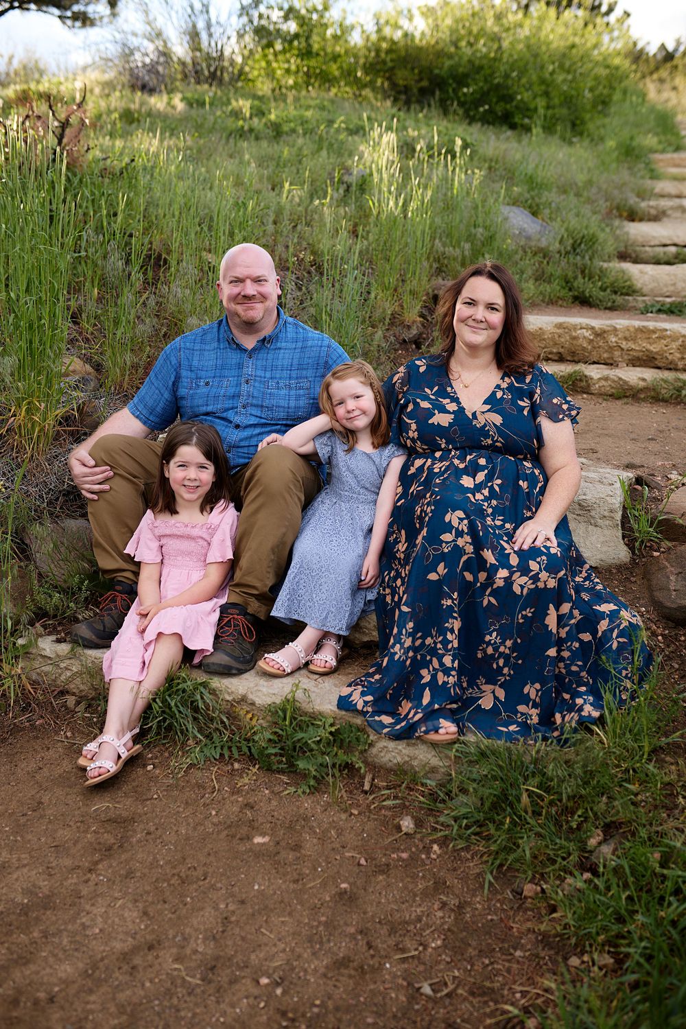 A smiling family of four with two young girls sits together on rustic stone steps surrounded by green grass on a sunny hillside.