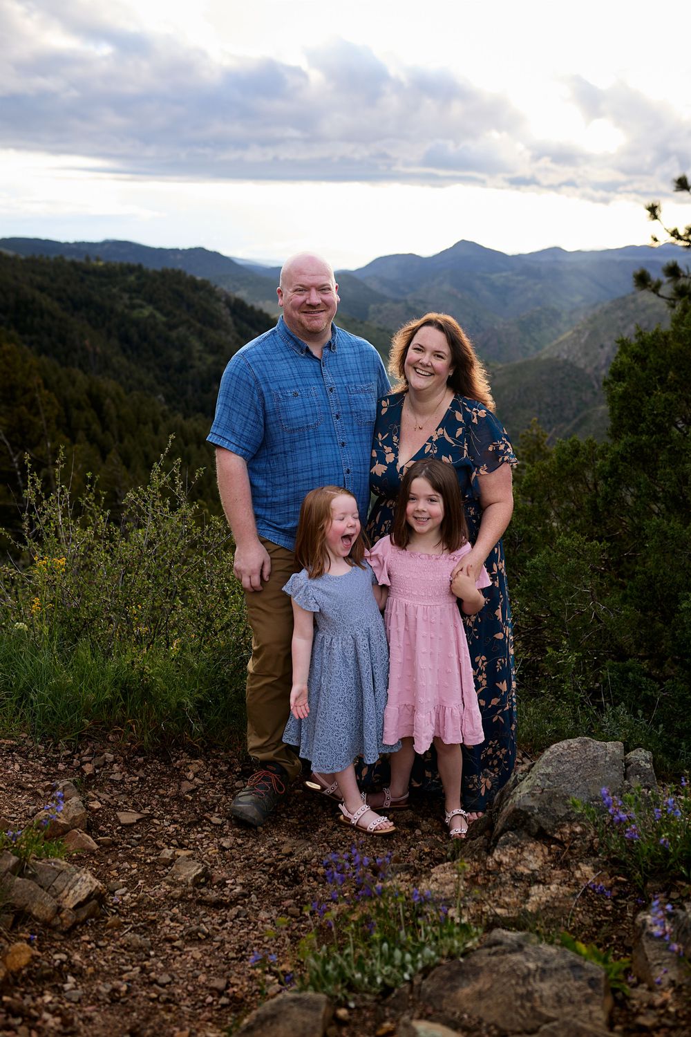 Family of four posing on a rocky mountain overlook with wildflowers, two young girls in dresses, and forested peaks beneath a cloudy sky.
