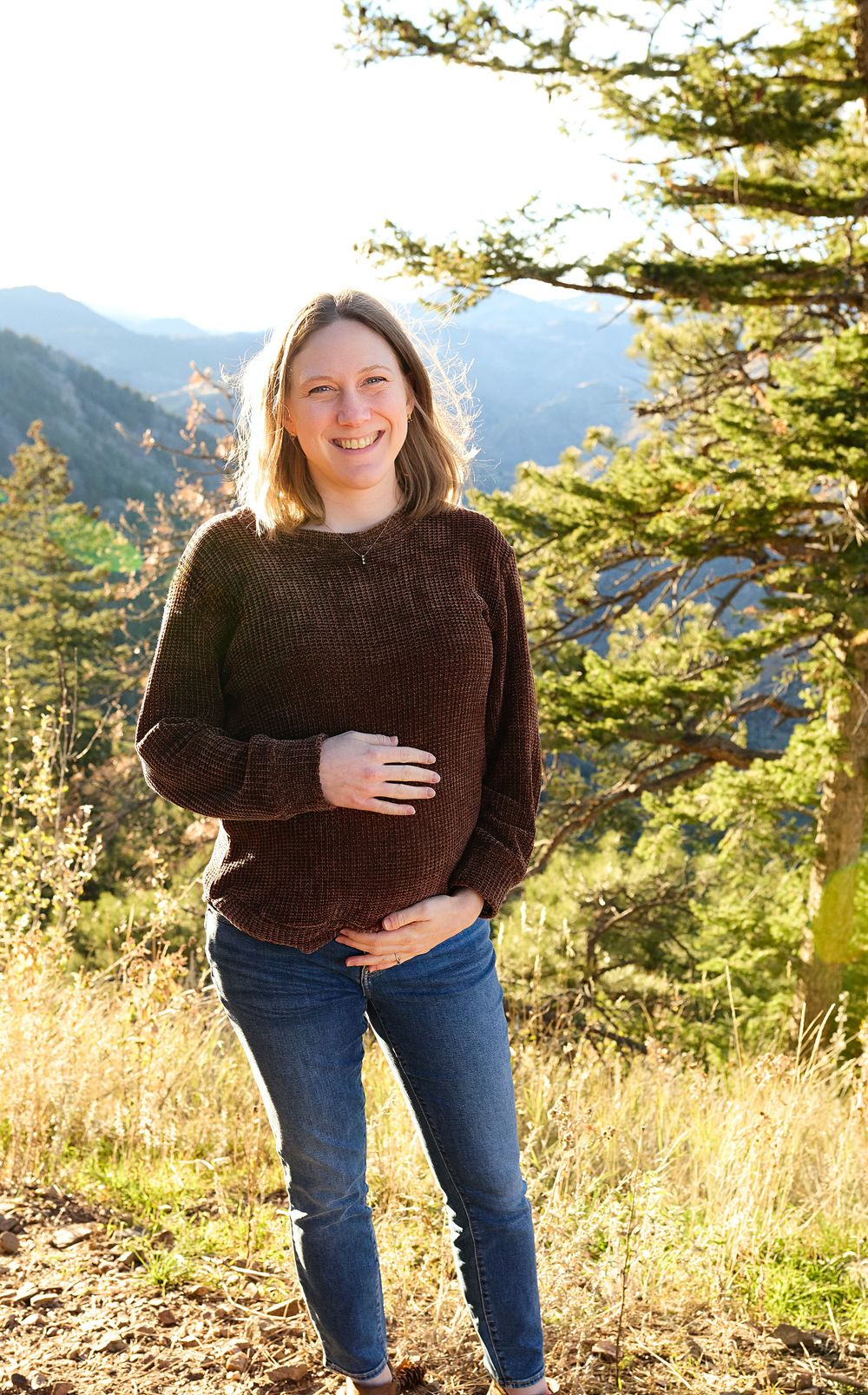 Smiling woman in a brown knit sweater and jeans stands with hands on her belly on a sunny mountain overlook with evergreen trees.