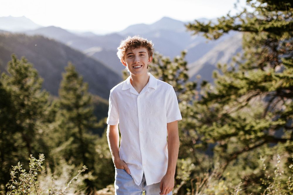 Smiling teen with curly hair in a white button-up shirt stands with hands in pockets among pine trees with sunlit mountains behind him.