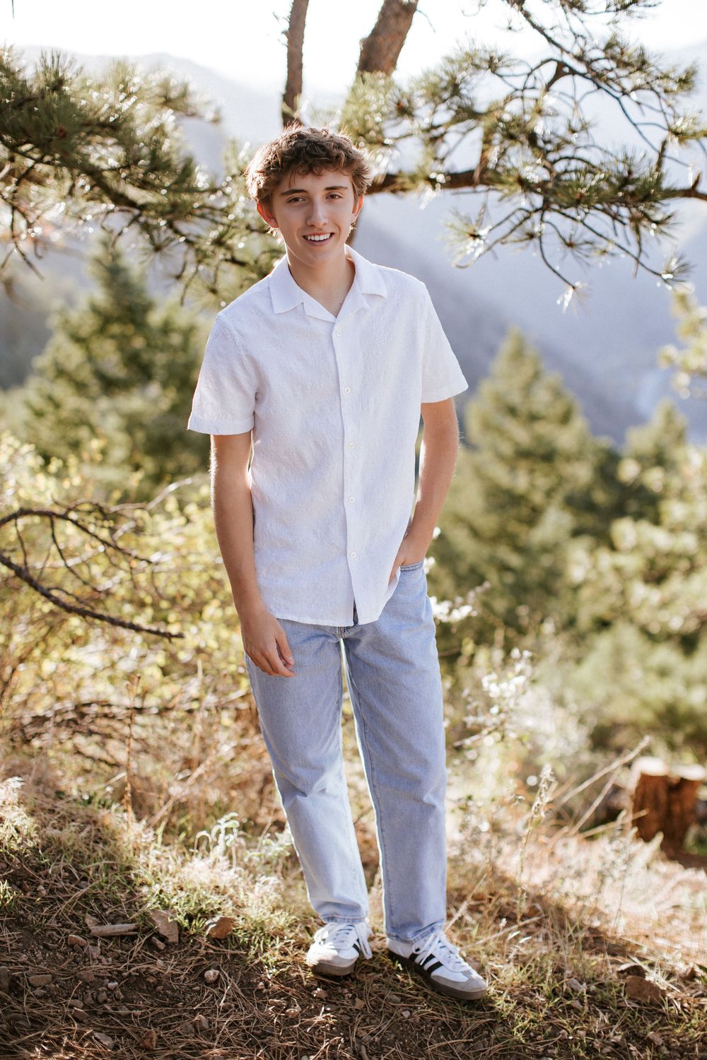 Smiling teenage boy in a white button-up shirt and light blue pants stands outdoors among pine trees with mountains in the background.