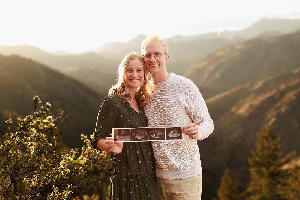 Smiling couple holding ultrasound photos at a scenic mountain overlook during golden hour, announcing their pregnancy.