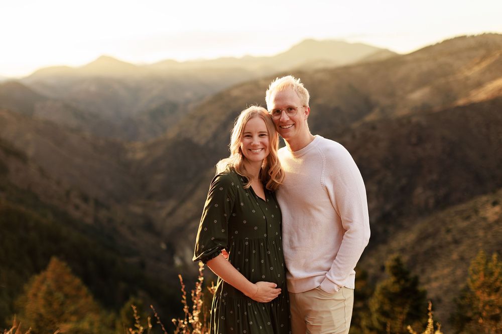 Smiling couple embracing in golden sunset light with rolling mountains and evergreen trees behind them, the woman in a green floral dress.