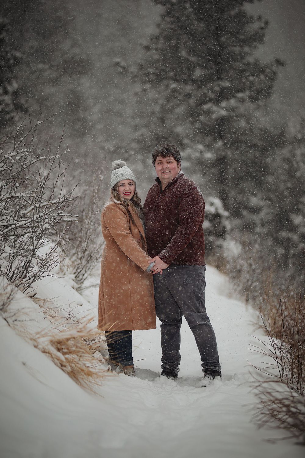 Smiling couple in winter coats holding hands on a snowy forest trail while snow falls around them.