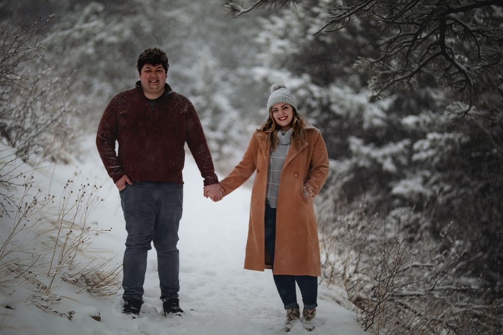 Smiling couple holding hands on a snowy forest path, he in a burgundy sweater and she in a camel coat and knit beanie.