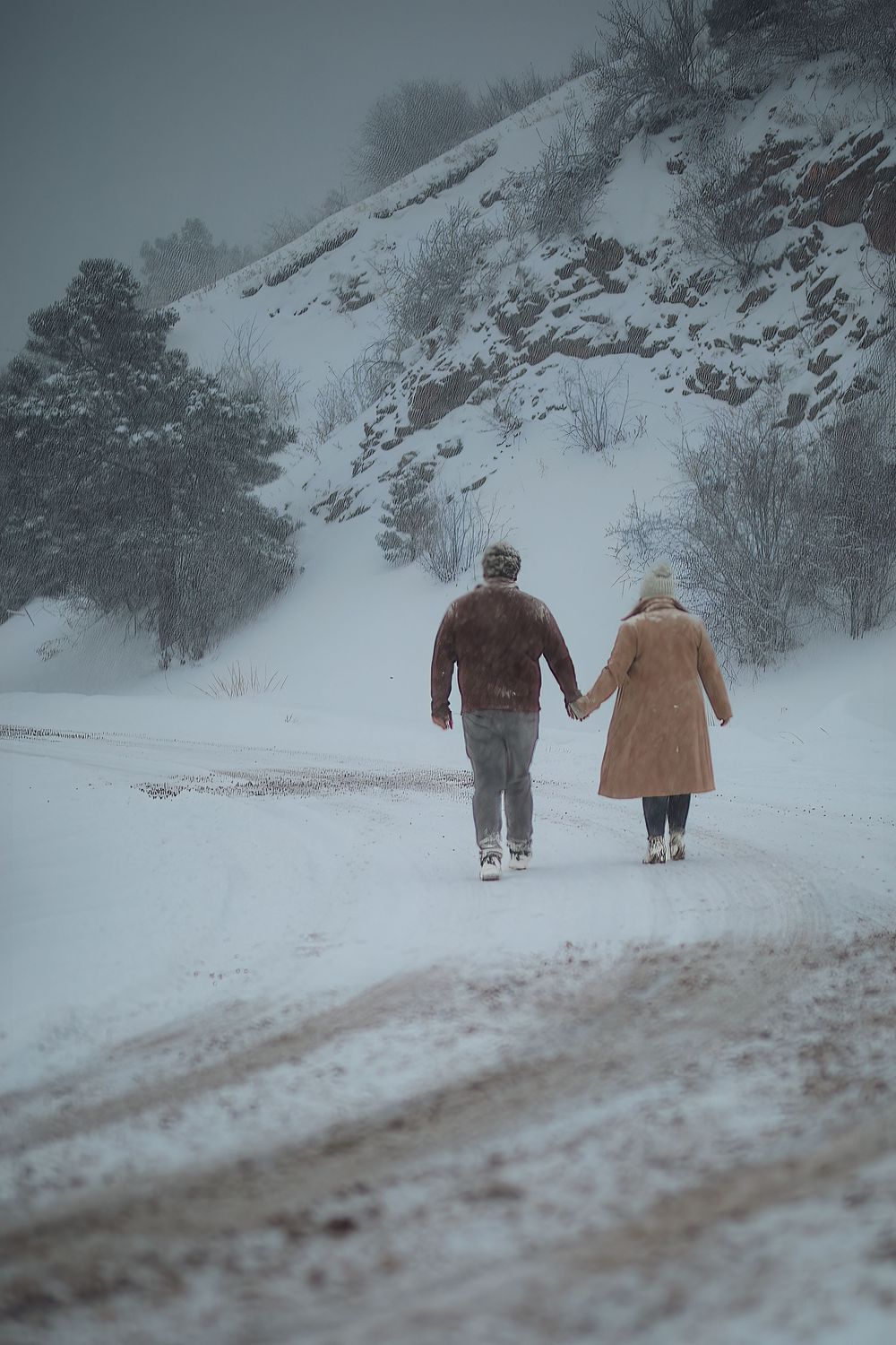 Couple holding hands walking along a snowy mountain road past a rocky hillside and evergreen trees during a winter snowfall.