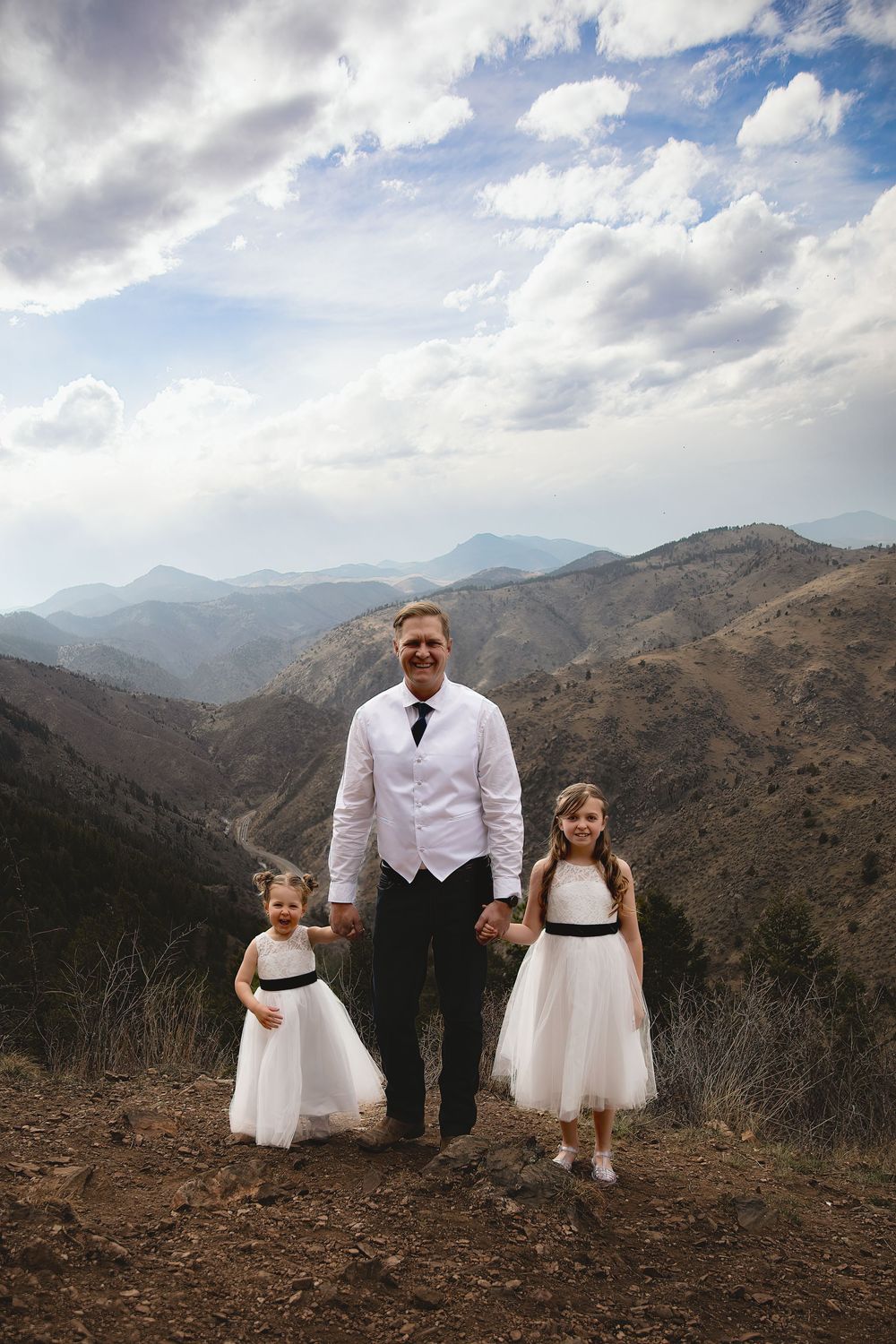 A man in a white shirt and black tie holds hands with two young girls in white dresses on a mountain overlook under a cloudy sky.