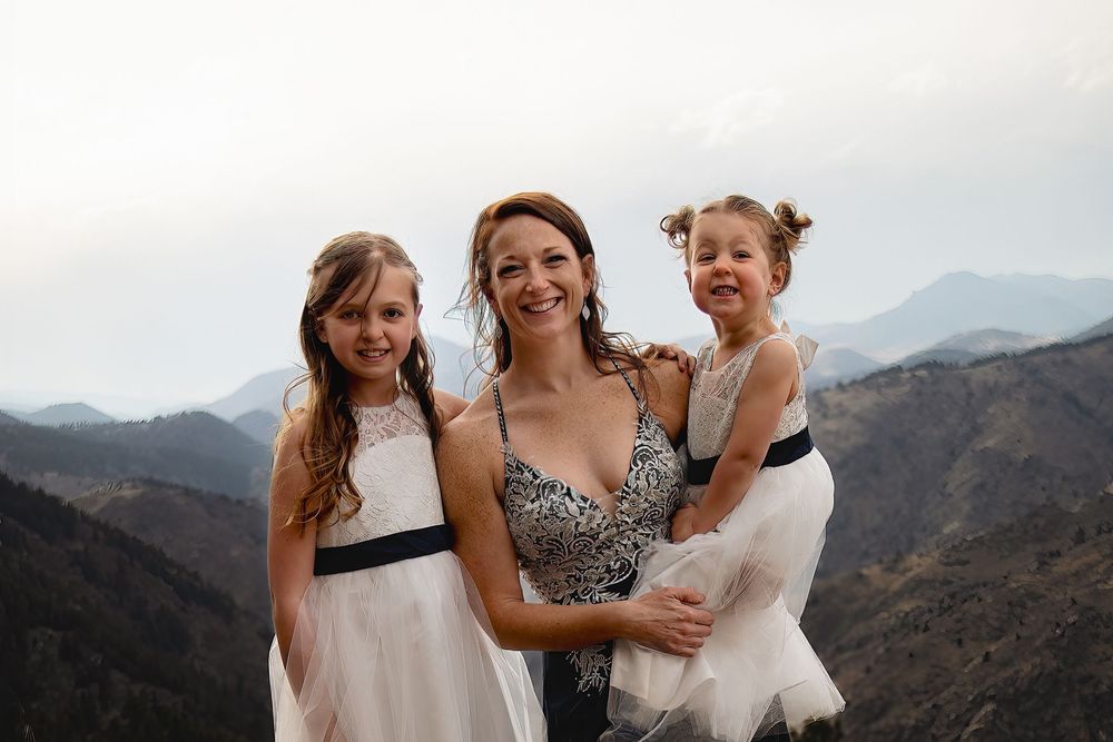 A smiling woman in a sparkly dress poses with two young girls in white dresses against a hazy mountain backdrop.