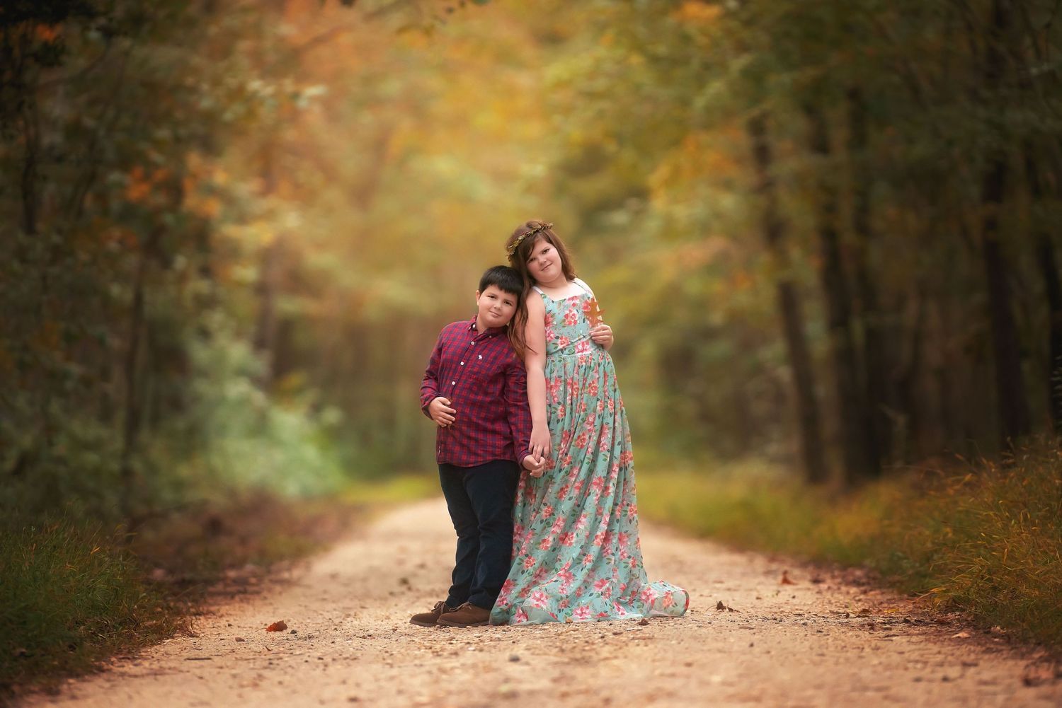A boy in a maroon shirt and a woman in a floral maxi dress hold hands on a dirt path in a sunlit autumn forest.