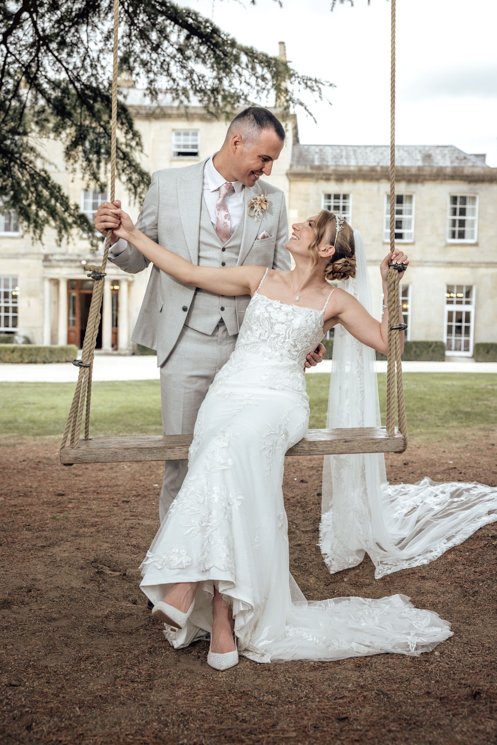 Bride in a lace wedding gown sits on a rope swing smiling up at her groom in a gray suit, with a grand manor house behind them.
