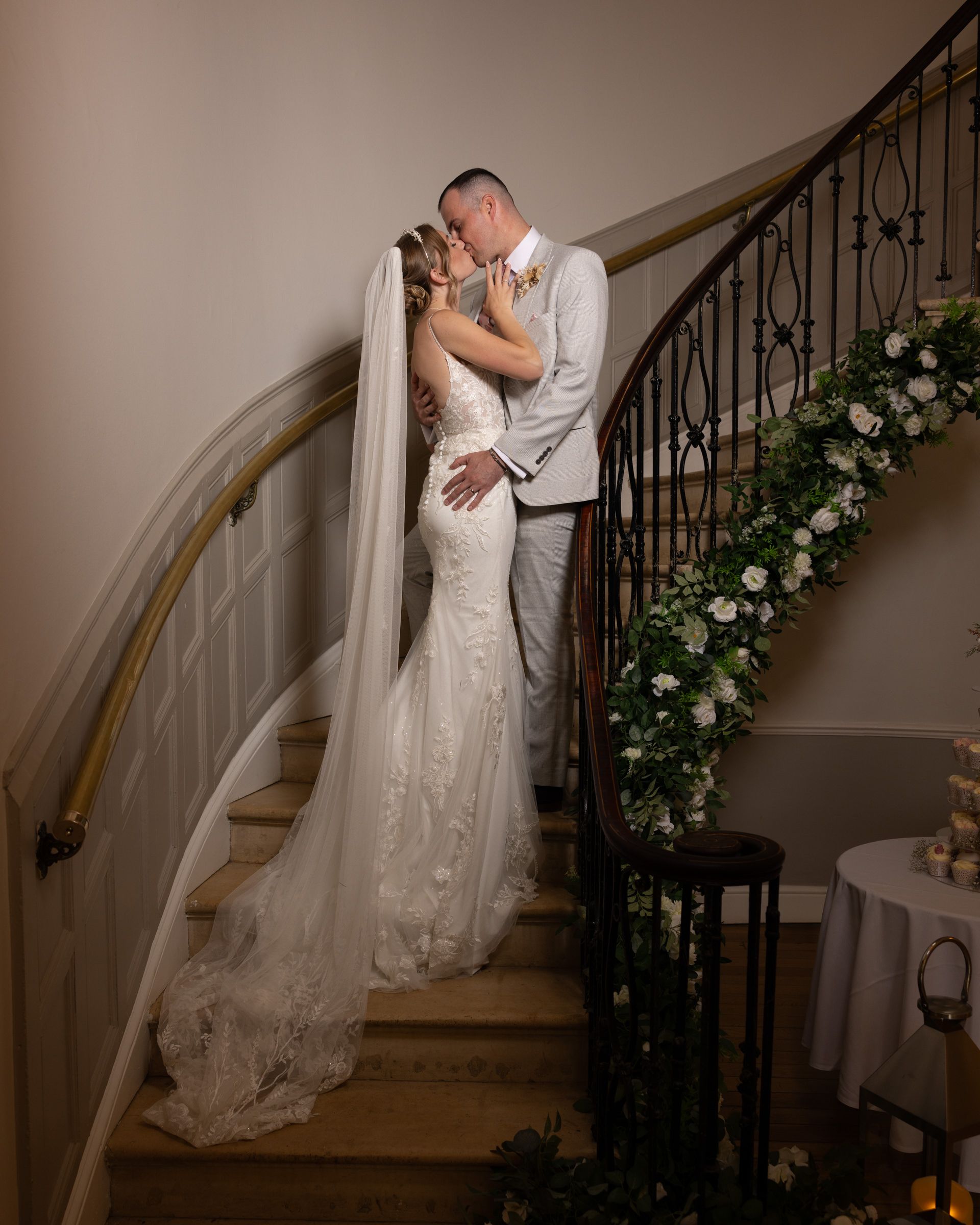 Bride in a lace gown with a long veil kisses her groom in a gray suit on an elegant curved staircase decorated with white floral garland.