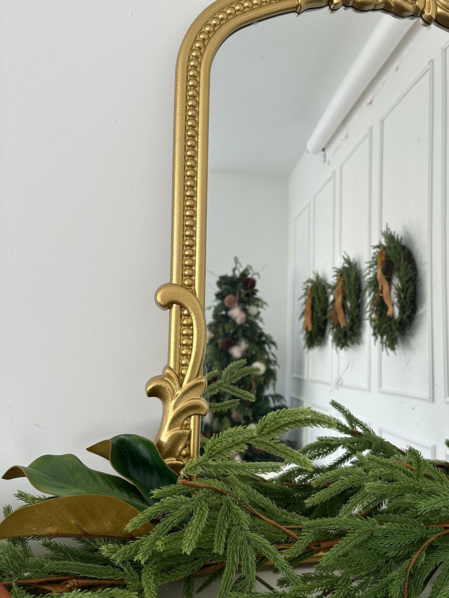 Gold ornate mirror reflecting a Christmas tree, with evergreen branches and magnolia leaves in the foreground.