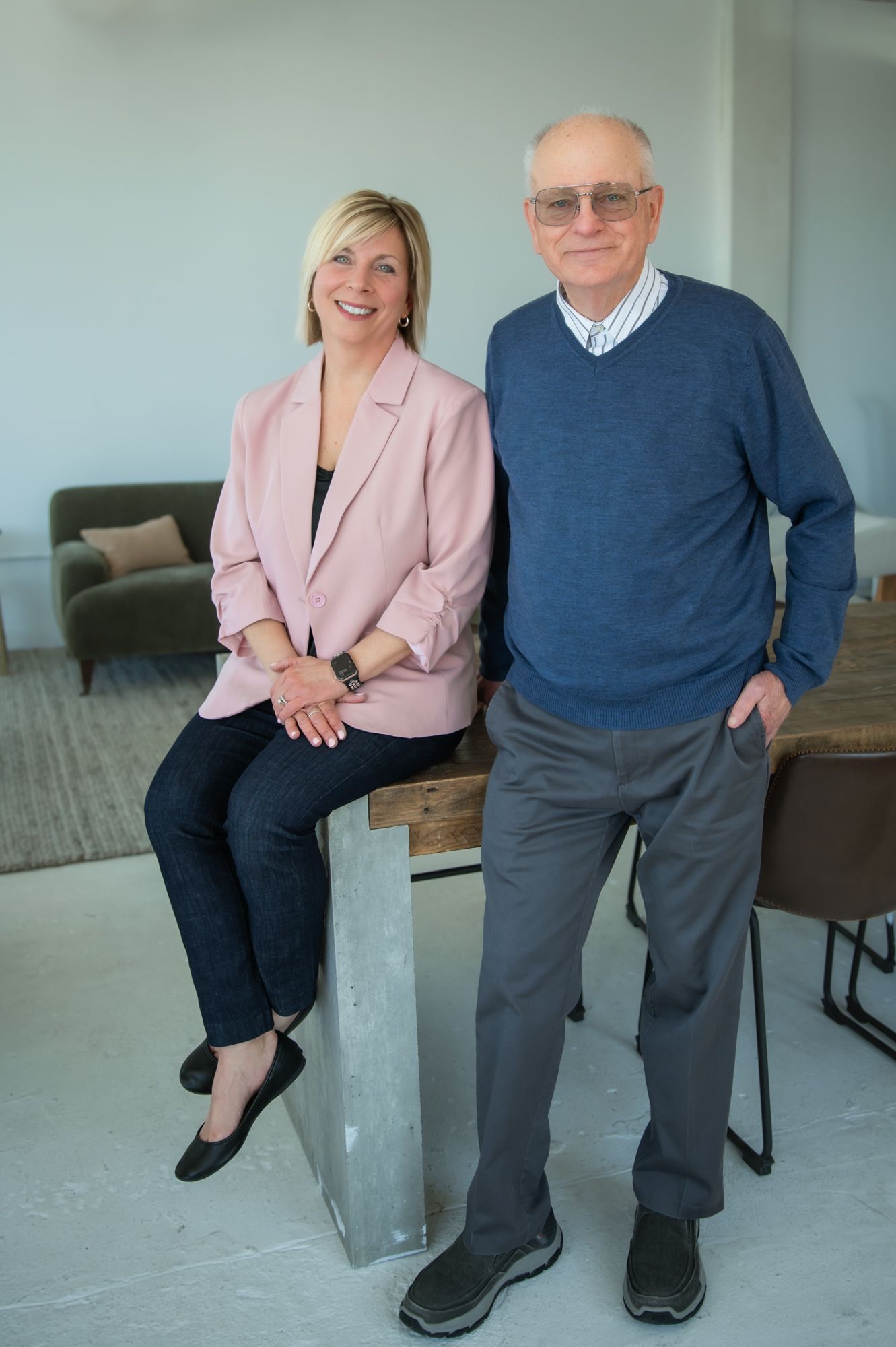 A smiling woman in a pink blazer sits beside an older man in a blue sweater in a modern bright interior space.